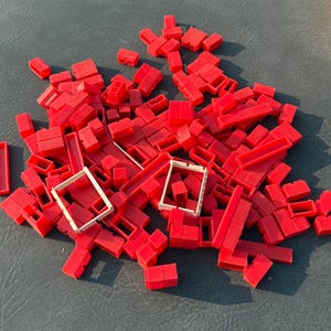 May include: A large collection of small red plastic building blocks in various rectangular shapes and sizes, scattered together with two small, off-white rectangular plastic frames positioned near the center of the pile.