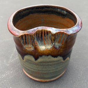 May include: A ceramic cup with a wide, rounded rim featuring a distinct notched indentation. The exterior displays a reactive glaze with blended tones of earthy brown, deep rust, and muted sage green, creating a layered, dripped appearance.
