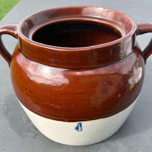 May include: A two-handled ceramic crock featuring a glossy dark brown glazed upper section and a matte off-white lower section. The lower portion displays a small, stamped blue number 4. The vessel has a wide circular opening and curved handles.