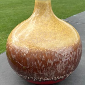 May include: A round ceramic vase with a narrow neck, featuring a glossy glaze that transitions from a mustard yellow at the top to a deep reddish-brown at the base, with a mottled, dripped texture effect throughout the surface of the vessel.