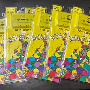 May include: Five identical Bobby Goldsboro presents Easter Egg Mornin' read-along book and cassette tape sets in yellow plastic packaging. Each set features a cartoon rabbit painting colorful Easter eggs on a bright yellow background.