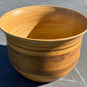May include: A round, wide-mouthed ceramic bowl featuring a warm, golden-brown glaze with darker horizontal bands and subtle, small circular indentations scattered across the exterior surface, showcasing a textured, hand-thrown appearance.