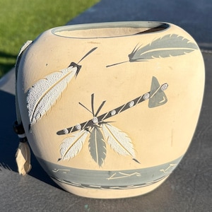 May include: A cream-colored ceramic vase with a rounded shape, featuring painted and textured feather motifs in gray and white. An arrow design with small white dots crosses the center, and a gray band encircles the base with a leather cord accent.