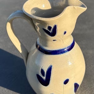 May include: A cream-colored ceramic pitcher featuring a glossy finish, a curved handle, and a wide pouring spout. The body and neck are decorated with hand-painted deep blue floral motifs and a solid blue horizontal band around the neck.