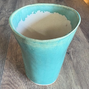 May include: A hand-thrown ceramic vessel with a flared rim and a tapered base. The exterior is finished in a vibrant turquoise glaze, while the interior reveals a contrasting off-white, unglazed clay surface that extends to the top edge.
