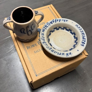 May include: A beige ceramic two-handled mug with blue lettering and a matching beige ceramic bowl featuring a blue checkerboard pattern and the alphabet and numbers zero through nine printed in blue around the rim, resting on a cardboard box.
