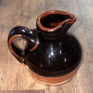 May include: A small ceramic pitcher featuring a rounded body and a curved handle. The upper portion is finished in a glossy, dark brown glaze with subtle speckling, while the base transitions into a warm, earthy terracotta orange tone.