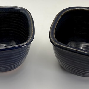 May include: Two deep, square-shaped ceramic bowls with rounded corners and a dark navy blue glaze. The exterior of each bowl features a series of horizontal ridges, while the interior is smooth and glossy with a deep blue finish.