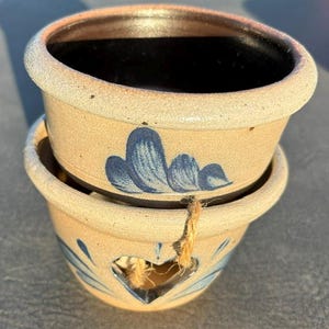 May include: Two stacked ceramic bowls with a speckled beige exterior and a smooth black interior. The top bowl features a blue painted floral design, while the bottom bowl has a heart-shaped cutout with a piece of brown twine tied through it.