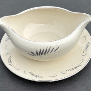 May include: A cream-colored gravy boat with an attached saucer, featuring a silver rim and a repeating gray fern-like leaf pattern. The gravy boat has a wide, curved pouring spout and a smooth, glossy finish on both the vessel and the base.