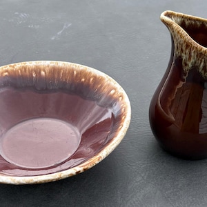 May include: A ceramic bowl and a matching creamer pitcher, both featuring a deep brown glossy glaze with a distinctive cream-colored drip pattern along the upper rims. The pitcher has a curved handle and a pouring spout, while the bowl is round.