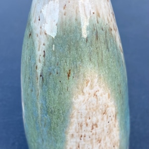 May include: A ceramic bud vase with a tapered neck and a rounded body. The glaze features a blend of sage green, cream, and white tones with brown speckles throughout. The rim is finished with a dark brown edge, and the base is unglazed.