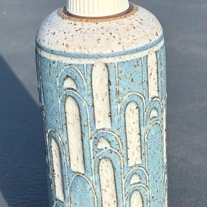 May include: A cylindrical ceramic soap dispenser featuring a speckled cream base with a textured blue arch pattern. The vessel is topped with a white plastic pump mechanism. The surface displays a repeating design of raised vertical arches.