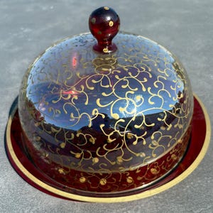 May include: A deep red glass cloche with a matching circular base, featuring a gold-painted swirling vine pattern across the dome. The lid is topped with a spherical knob, and the base is finished with a thin gold rim along the outer edge.