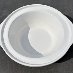 May include: A round white ceramic serving bowl with integrated side handles. The bowl features a smooth, glossy finish and a wide, flat rim that extends outward from the central basin, creating a clean and functional design for serving food.