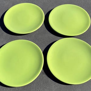 May include: A set of four round, lime green plates arranged in a two-by-two grid. Each plate features a smooth, matte surface with a simple, circular design and a slightly raised edge, providing a uniform appearance across all four pieces.