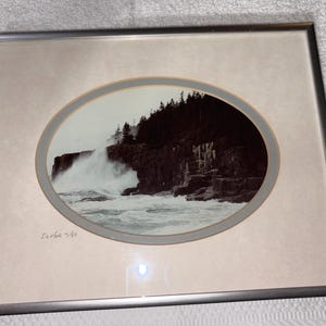 May include: A framed black and white photograph featuring an oval-shaped landscape of crashing ocean waves against a rocky cliffside with pine trees. The print is matted in a light beige border and signed with the text Dover 14/50 in the corner.
