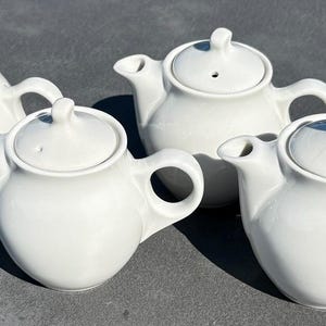 May include: A set of four identical white ceramic teapots, each featuring a rounded body, a curved spout, a circular handle, and a matching lid with a small knob handle and a single steam vent hole, arranged in a group on a flat surface.