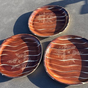 May include: Three round plates with a dark brown wood-grain finish, featuring horizontal wavy white lines and a central etched floral design. Each plate has a smooth, glossy surface and a slightly raised edge, showcasing a consistent pattern.