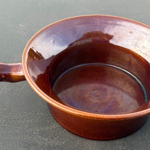 May include: A round, dark brown ceramic bowl with a single, thick, integrated handle extending from one side. The bowl features a smooth, glossy, reflective glaze finish, a wide rim, and a deep, circular basin with a flat bottom surface.