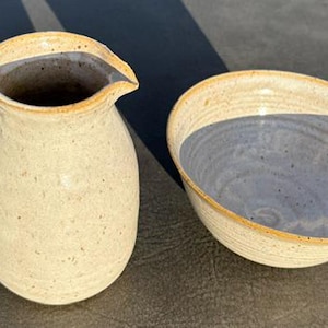 May include: A ceramic pitcher and a matching bowl featuring a speckled cream-colored glaze with light brown rims. The pitcher has a curved handle and a pouring spout, while the bowl has a wide, open design with a smooth, light gray interior surface.