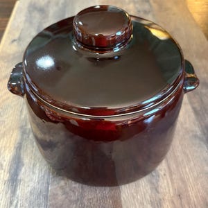 May include: A dark brown ceramic pot with a matching lid and two small side handles. The pot features a smooth, glossy finish that reflects light, creating a uniform appearance across the rounded body and the circular lid with a central knob.