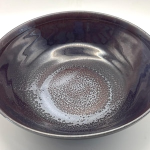 May include: A round ceramic bowl with a glossy finish, featuring a deep dark purple and black color palette. The interior surface displays a textured, speckled pattern in the center that transitions into a smooth, dark glaze toward the rim.
