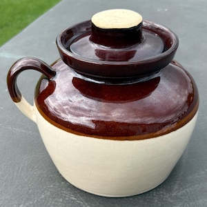 May include: A ceramic pot with a lid, featuring a two-tone design with a glossy dark brown glaze on the upper portion and lid, and a matte cream-colored finish on the lower body. The pot includes a single curved handle on the left side.