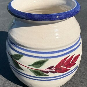 May include: A cream-colored ceramic pot with a wide, rounded body and a thick, dark blue rim. The exterior features two horizontal blue stripes encircling the middle, with a painted botanical design of green leaves and red foliage in between.