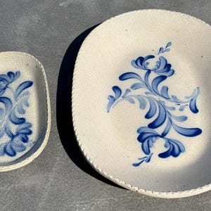 May include: Two oval-shaped ceramic dishes with a speckled off-white glaze and a textured rope-like edge. Each dish features a hand-painted blue floral motif with swirling leaves and petals centered on the interior surface of the pottery.