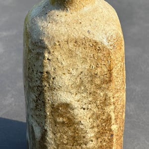May include: A tall, cylindrical ceramic vase with a textured, speckled tan and brown surface. The vessel features a narrow neck, a flared rim, and a faceted body with visible clay grain, showcasing a natural, earthy finish throughout the piece.