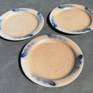 May include: Three round ceramic plates with a speckled beige finish and a wide rim. Each plate features a hand-painted cobalt blue leaf pattern arranged in a circular motif along the outer edge, creating a consistent design across all three pieces.