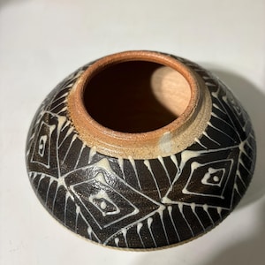 May include: A low-profile, round ceramic bowl with a wide opening and a thick, unglazed tan rim. The exterior features a dark brown, textured glaze decorated with a repeating pattern of white, hand-painted geometric diamond shapes and lines.