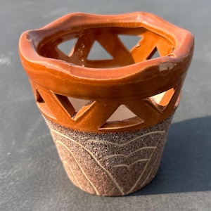 May include: A ceramic vessel featuring a glossy, smooth orange upper section with triangular cutouts and a wavy rim. The lower portion has a textured, matte brown finish with etched, light-colored geometric line patterns encircling the base.