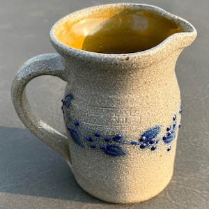 May include: A stoneware pitcher with a speckled beige exterior and a smooth, glossy mustard-yellow interior. The pitcher features a blue painted vine motif with leaves and berries, and a stamped mark reading "JASON HILL KILNS, MASON, N.H.".