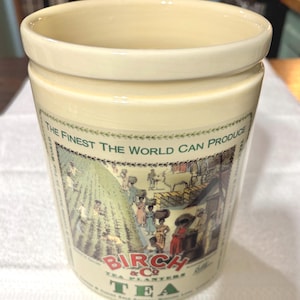 May include: A cream-colored ceramic canister featuring a decorative illustration of a tea plantation with workers and an ox. The text reads: THE FINEST THE WORLD CAN PRODUCE, BIRCH & CO TEA PLANTERS, and TEA. The cylindrical container has a smooth finish.