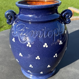 May include: A deep blue ceramic jar with two side handles and a wide opening. The exterior features the embossed word Rumtopf in cursive script and a repeating pattern of small white three-dot clusters. The interior rim is a warm brown color.