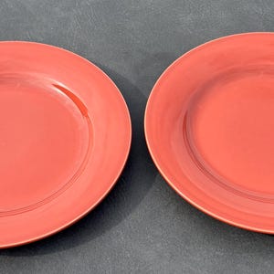 May include: Two round, solid coral-red dinner plates with a smooth, glossy finish. Each plate features a slightly raised rim and a flat center, showcasing a consistent, vibrant color throughout the surface of the ceramic dinnerware set.