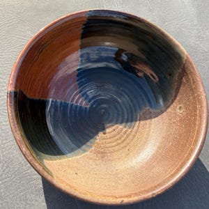 May include: A round ceramic bowl featuring a two-tone glaze with a deep navy blue section contrasting against a warm, speckled terracotta brown base. The interior surface displays visible concentric throwing rings and a smooth, glossy finish.