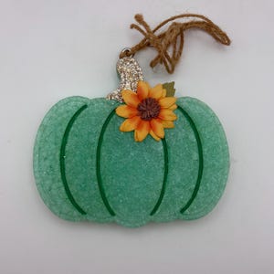 May include: A mint green pumpkin-shaped ornament with a textured surface and a glittery silver stem. A single orange and yellow sunflower with a brown center is attached to the top. A brown twine loop is tied to the stem for hanging purposes.
