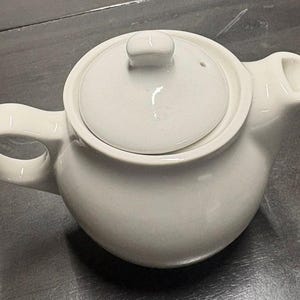 May include: A solid white ceramic teapot featuring a rounded body, a curved handle on the left side, a short spout on the right side, and a matching circular lid with a small knob handle centered on top. The teapot has a smooth, glossy finish.