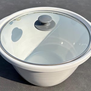 May include: A round white ceramic cooking pot with integrated side handles and a clear glass lid. The lid features a metallic rim and a solid gray circular knob handle centered on top. The pot has a smooth, glossy finish and a deep basin.
