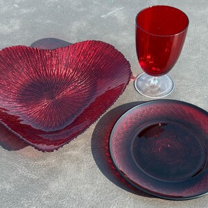 May include: A set of deep red glassware consisting of a heart-shaped bowl with a textured sunburst pattern, a round plate featuring a cross-hatched grid design, and a tall, smooth-surfaced stemmed goblet with a clear glass base.