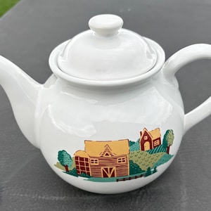 May include: A white ceramic teapot with a lid, featuring a colorful graphic print of a red barn, a smaller house, and green trees. The teapot has a curved handle and a spout, with a smooth, glossy finish and a rounded body shape.