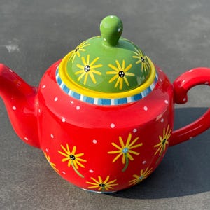 May include: A bright red ceramic teapot featuring a green lid decorated with yellow daisy-like flowers. The body of the teapot is adorned with small white polka dots and larger yellow flowers, finished with a blue and white striped rim detail.
