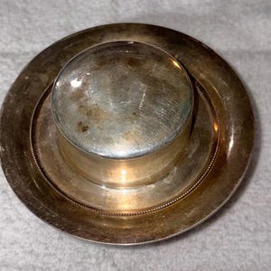 May include: A circular silver-toned metal container with a smooth, domed lid and a wide, flat rim. The surface exhibits a polished finish with visible signs of wear and light tarnishing, featuring a beaded texture along the inner edge.