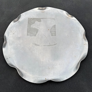May include: A round, silver-colored metal tray with a scalloped, wavy edge. The center of the tray features a lightly etched, line-drawn illustration of a stylized figure standing in profile, set against a rectangular, textured background.