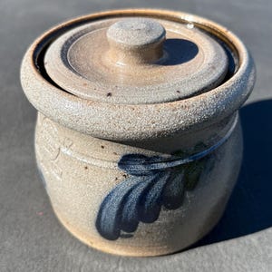 May include: A beige stoneware crock with a matching lid, featuring a speckled texture and a hand-painted blue leaf design on the front. The cylindrical container has a thick, rounded rim and a small knob handle on the center of the lid.