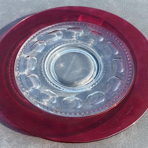 May include: A round glass plate featuring a deep burgundy red outer rim and a clear center section. The clear portion is decorated with a ring of embossed oval indentations and a delicate beaded texture border surrounding the central circular area.