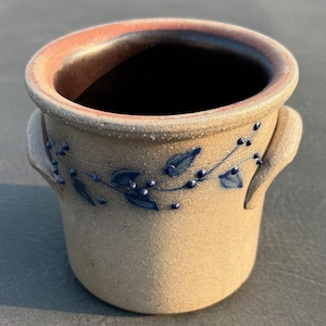 May include: A cylindrical stoneware crock with two side handles, featuring a textured tan exterior and a dark brown interior. The exterior is decorated with a hand-painted blue vine pattern consisting of small leaves and dots encircling the body.